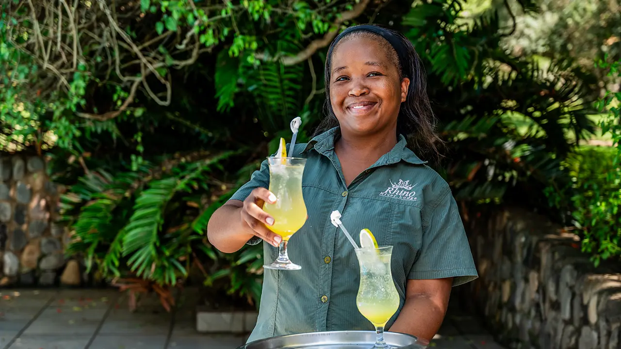 A smiling employee at Rhino River Lodge stands outdoors in a khaki uniform, holding a silver tray with refreshing drinks for guests to enjoy.