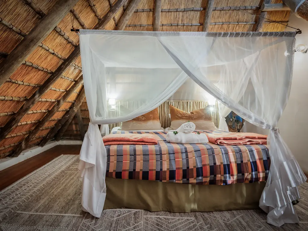 A view of the upstairs bedroom inside a luxury safari Cottage at Rhino River Lodge, showcasing a neatly made double bed tucked beneath the sloping thatch-roof ceiling.