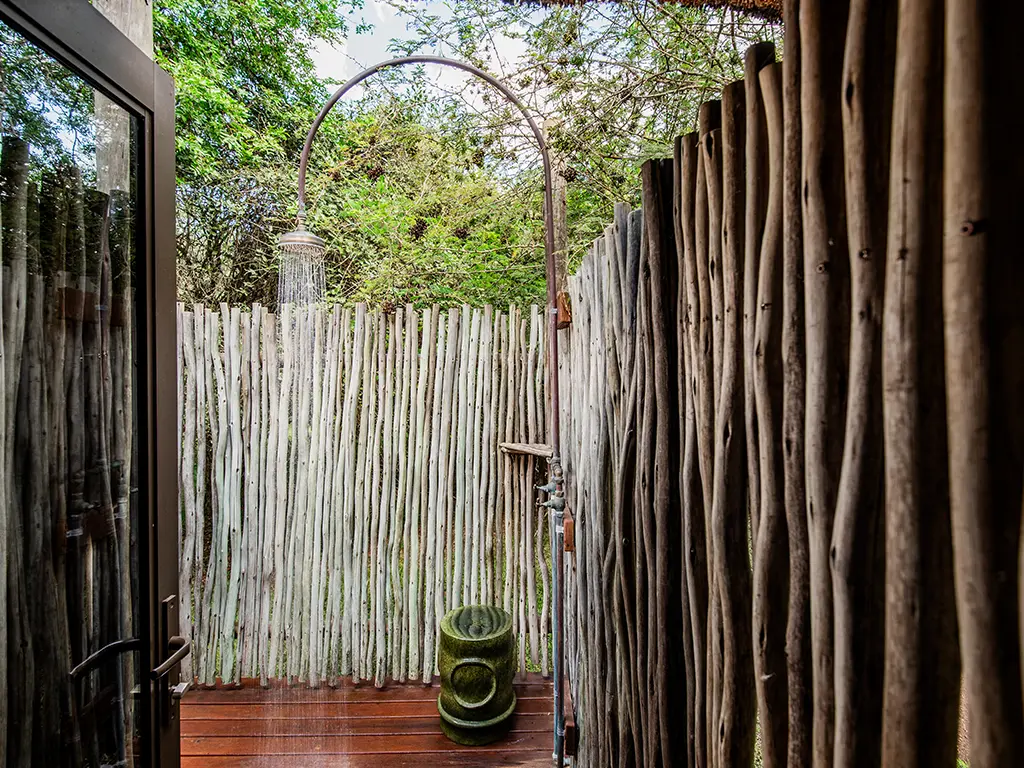 A refreshing outdoor shower built on a wooden deck and enclosed by a rustic reed privacy screen at a luxury safari Cottage at Rhino River Lodge, surrounded by lush green bushveld trees.
