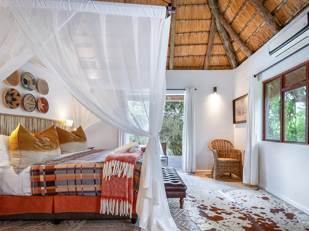 A side view inside a luxury safari Cottage bedroom at Rhino River Lodge, showing a comfortable double bed draped in white mosquito netting, positioned next to large glass doors that look out onto the lush green bushveld.