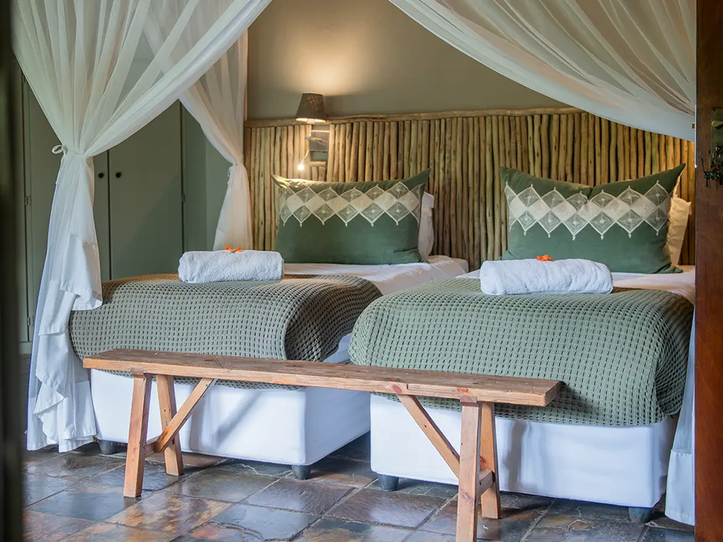 A close-up view of two comfortable beds neatly made with white linen and decorative green cushions inside a luxury safari Deluxe Room at Rhino River Lodge.