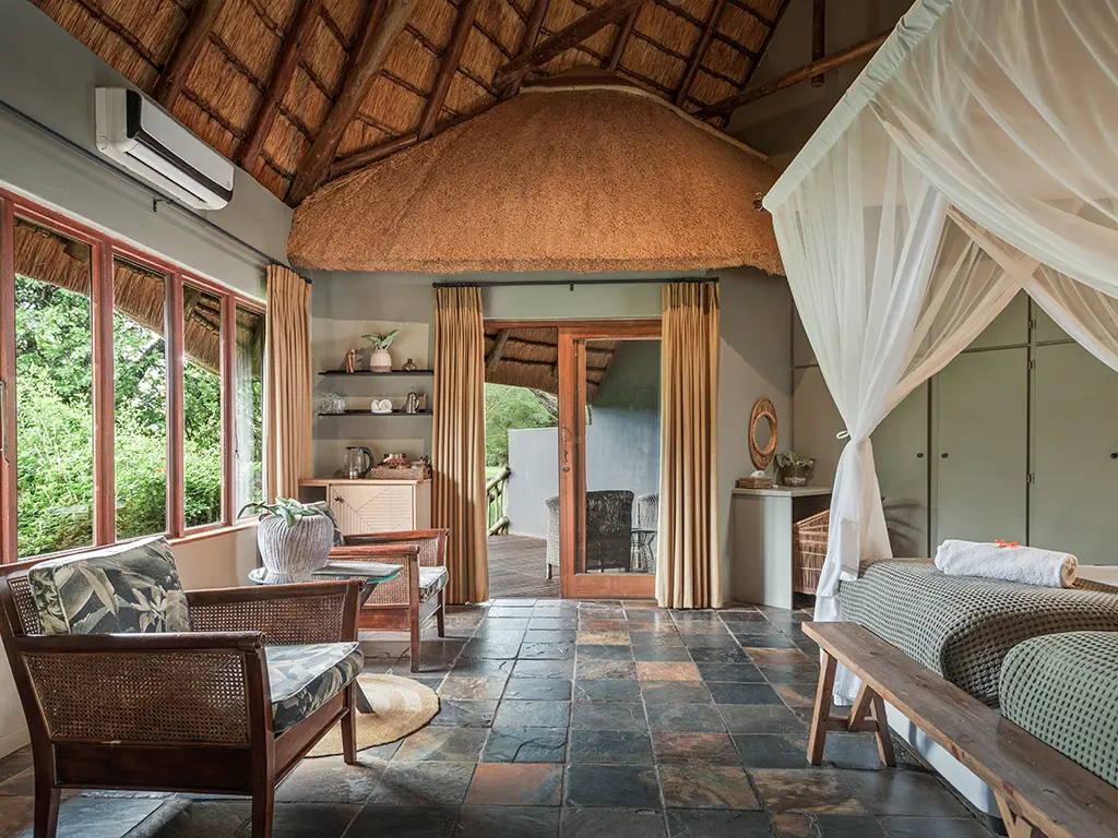 A wide interior view of a luxury safari Deluxe Room at Rhino River Lodge, looking toward the entrance, featuring exposed wooden ceiling beams, a tiled floor with a woven rug, a writing desk.