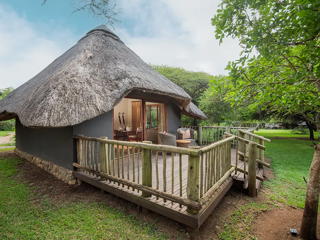 An outdoor view from a luxury safari Deluxe Room at Rhino River Lodge, looking out through large glass doors onto a private wooden deck surrounded by lush bushveld trees.