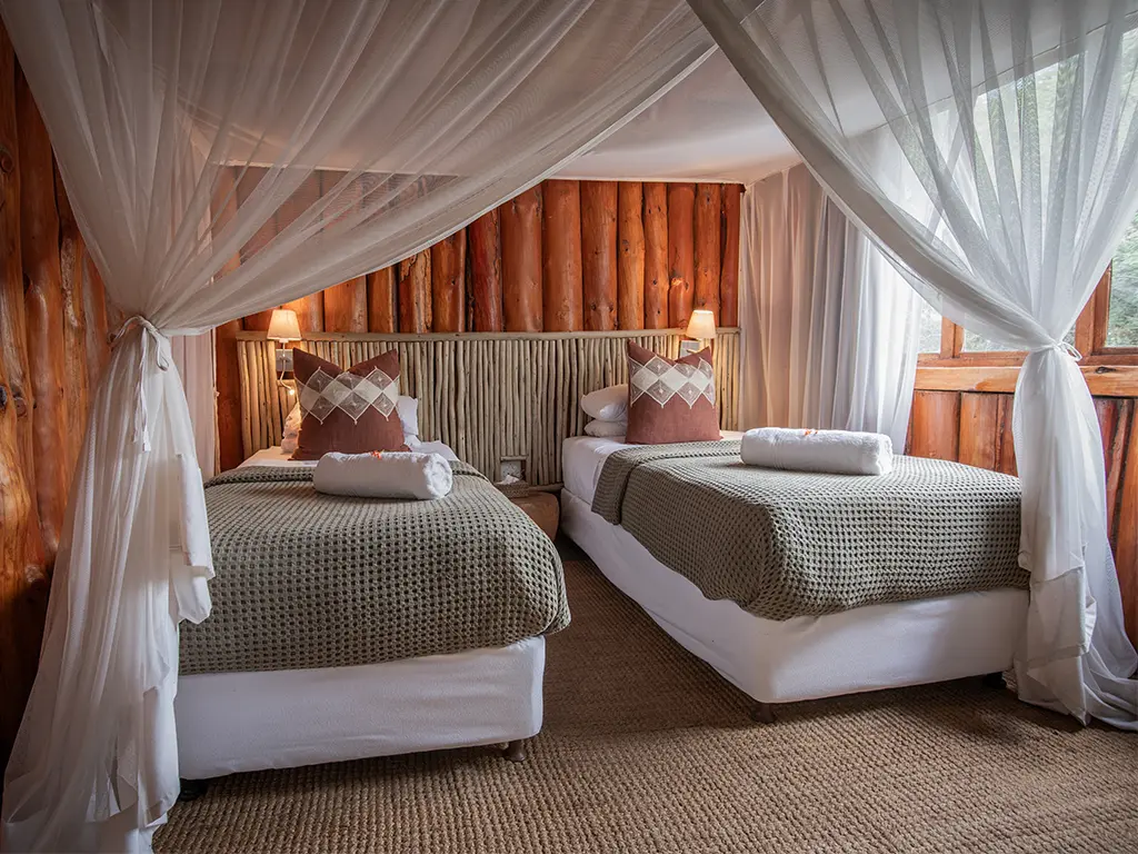 One of the bedrooms inside a family loft chalet at Rhino River Lodge, showcasing two single beds made up with white linen and brown throw cushions, set against a feature wall under ambient reading lights.