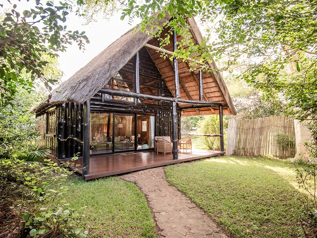 An exterior view of a double-story Family Loft Chalet at Rhino River Lodge, showcasing a large thatched roof, and an outdoor wooden deck surrounded by lush bushveld trees.