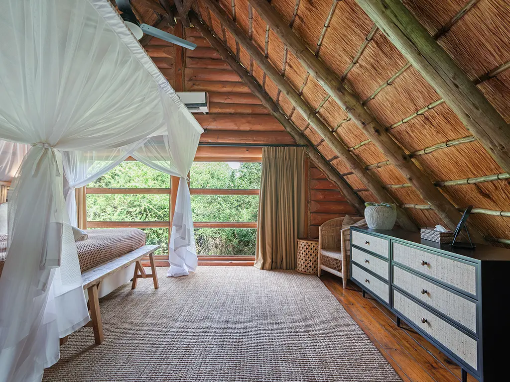 A side angle view of the upstairs loft bedroom in a Family Loft Chalet at Rhino River Lodge, showing a beautifully made bed under an exposed thatched roof ceiling with elegant wooden decor.