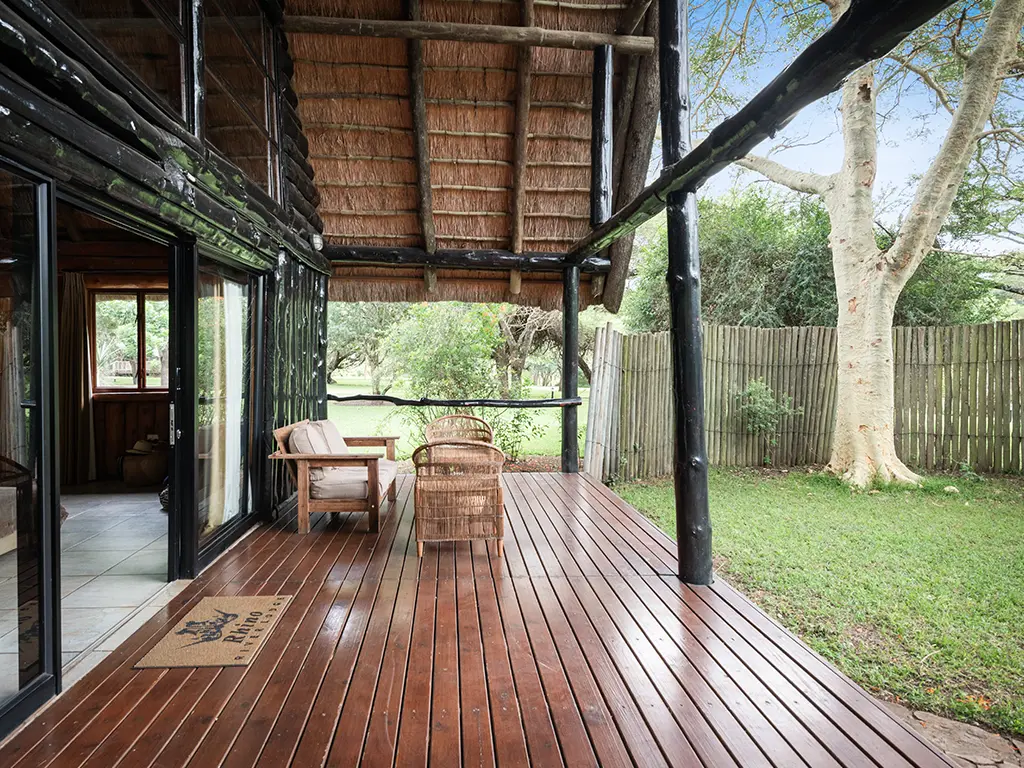 An exterior view of the entrance and large private wooden deck of a Family Loft Chalet at Rhino River Lodge, featuring outdoor seating surrounded by lush green bushveld trees.