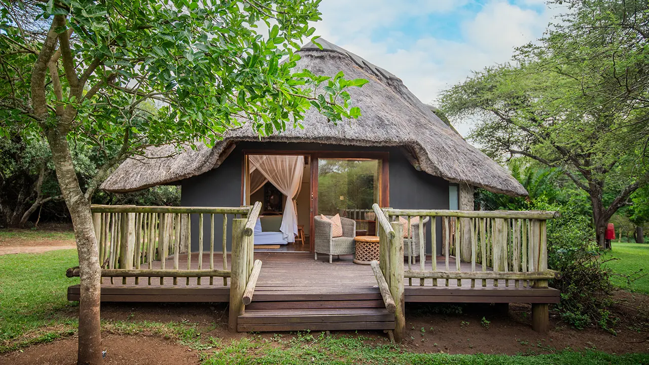 A thatched-roof luxury safari chalet with an open wooden deck and woven chairs, surrounded by lush green trees at Rhino River Lodge.