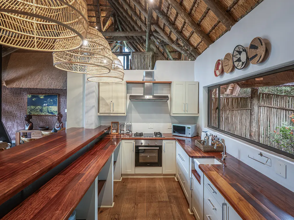 Interior view of the fully equipped kitchen inside the Homestead at Rhino River Lodge, featuring modern wooden cabinetry, a central island counter, and stainless steel appliances beneath a rustic thatched ceiling.