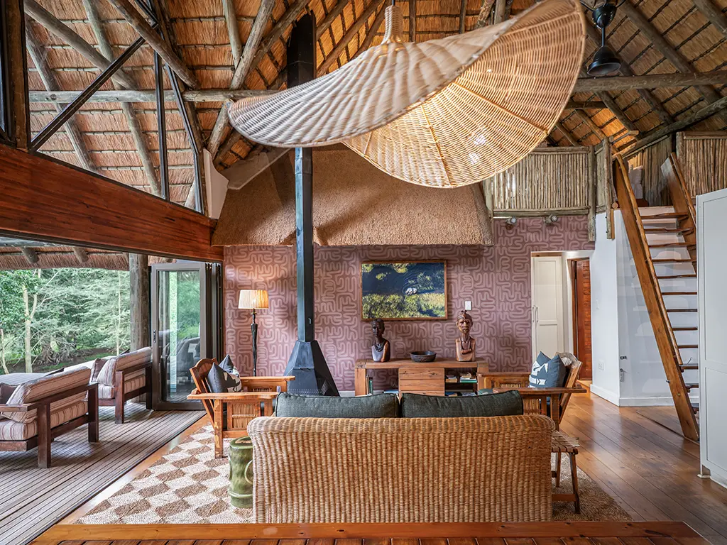 An interior view of the open-plan lounge inside the Homestead at Rhino River Lodge, captured from the dining area, showcasing comfortable plush sofas arranged around a coffee table, a beautiful painting hanging above a wooden cabinet adorned with statues, and large glass sliding doors that look out onto the lush outdoor greenery.