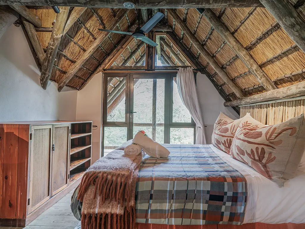 Interior view of the upstairs loft bedroom in the Homestead at Rhino River Lodge, featuring a double bed with a plaid and fringed blanket tucked under a steep, rustic thatched roof with exposed log beams, a black ceiling fan, and a wooden storage cabinet and shelving unit on the left next to glass doors.