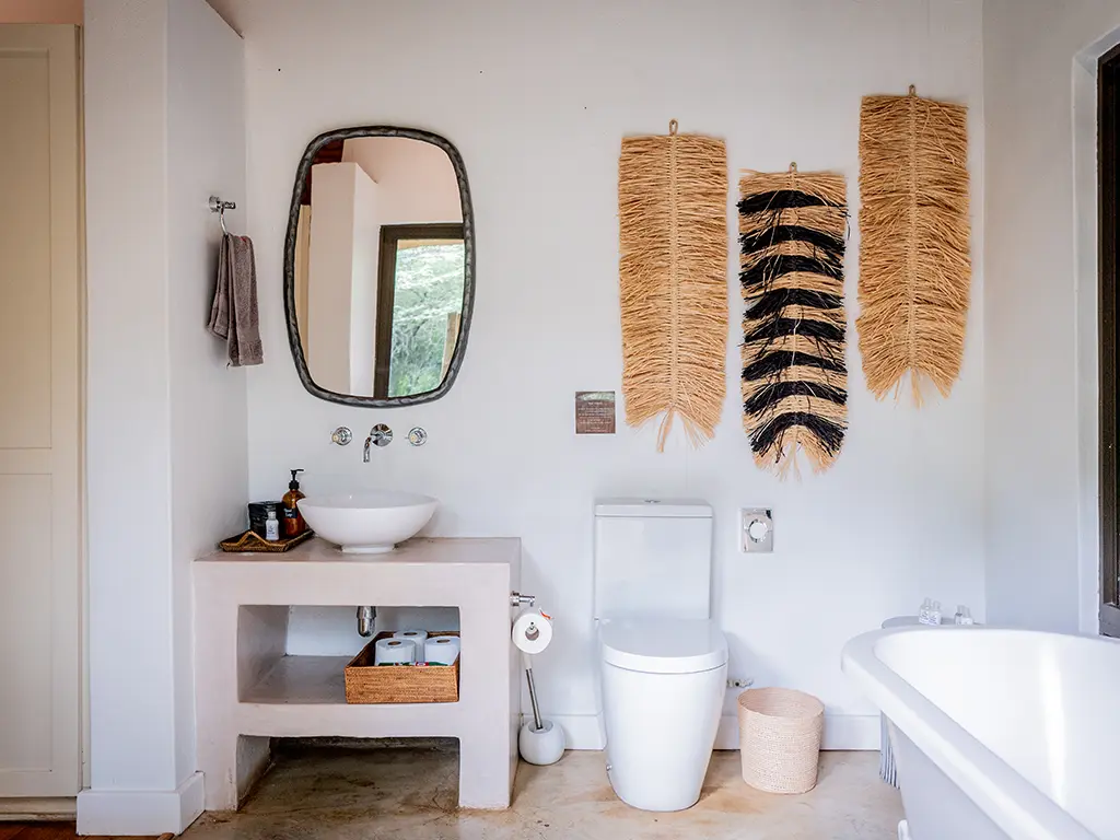 Interior view of a bathroom inside the Homestead at Rhino River Lodge, featuring a white vessel sink on a raised concrete vanity, a big mirror, a white toilet next to a woven bin, and three fringed wall hangings above it, with a white freestanding bathtub partially visible on the right.