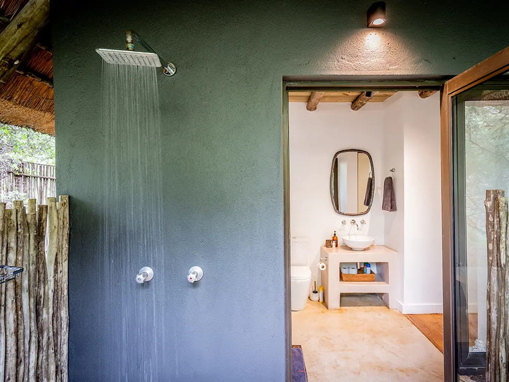 View from an outdoor shower looking into a bathroom at the Homestead at Rhino River Lodge, showing a portion of a white toilet on the concrete floors inside, with the outdoor shower area enclosed by rustic wooden sticks for privacy and surrounded by green trees.