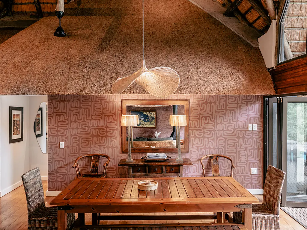 Interior view of the dining area inside the Homestead at Rhino River Lodge, featuring a large wooden dining table surrounded by woven chairs under a large woven pendant light, with a freestanding three-drawer wooden cabinet flanked by two tall lamps in the background.
