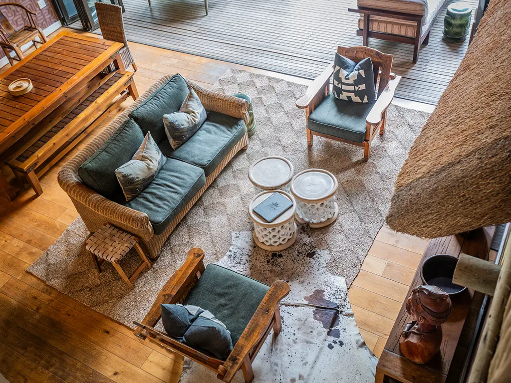 A high-angle view looking down from the loft into the open-plan living area of the Homestead at Rhino River Lodge, showing a dark green upholstered woven sofa and matching armchairs arranged around a cluster of three small textured round coffee tables on a large patterned rug, with a cowhide rug layered on top, wooden flooring, and a partial view of a wooden dining table setup and a thatched fireplace structure to the sides.