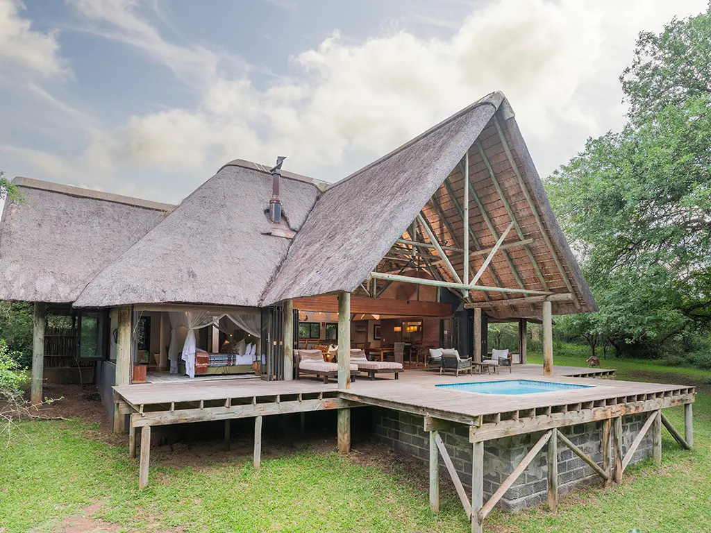 A high-angle, drone view looking at the Homestead at Rhino River Lodge, showing the double-story luxury safari villa nestled in thick African bush, featuring a steep thatched roof, private wooden deck with a swimming pool and outdoor seating, surrounded by lush green trees.