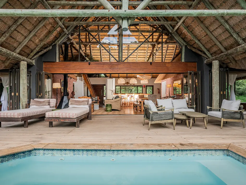 An outdoor view from the deck of the Homestead at Rhino River Lodge, looking across the spacious wooden patio with outdoor lounge seating toward the open stack-away doors that reveal the indoor open-plan dining and living area.