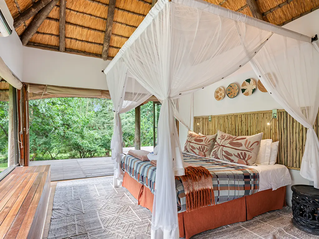 Interior view of a luxurious bedroom inside the Homestead at Rhino River Lodge, featuring a double bed draped in a white mosquito net canopy under a high thatched roof, with a rustic wooden headboard and a woven rug on the floor.