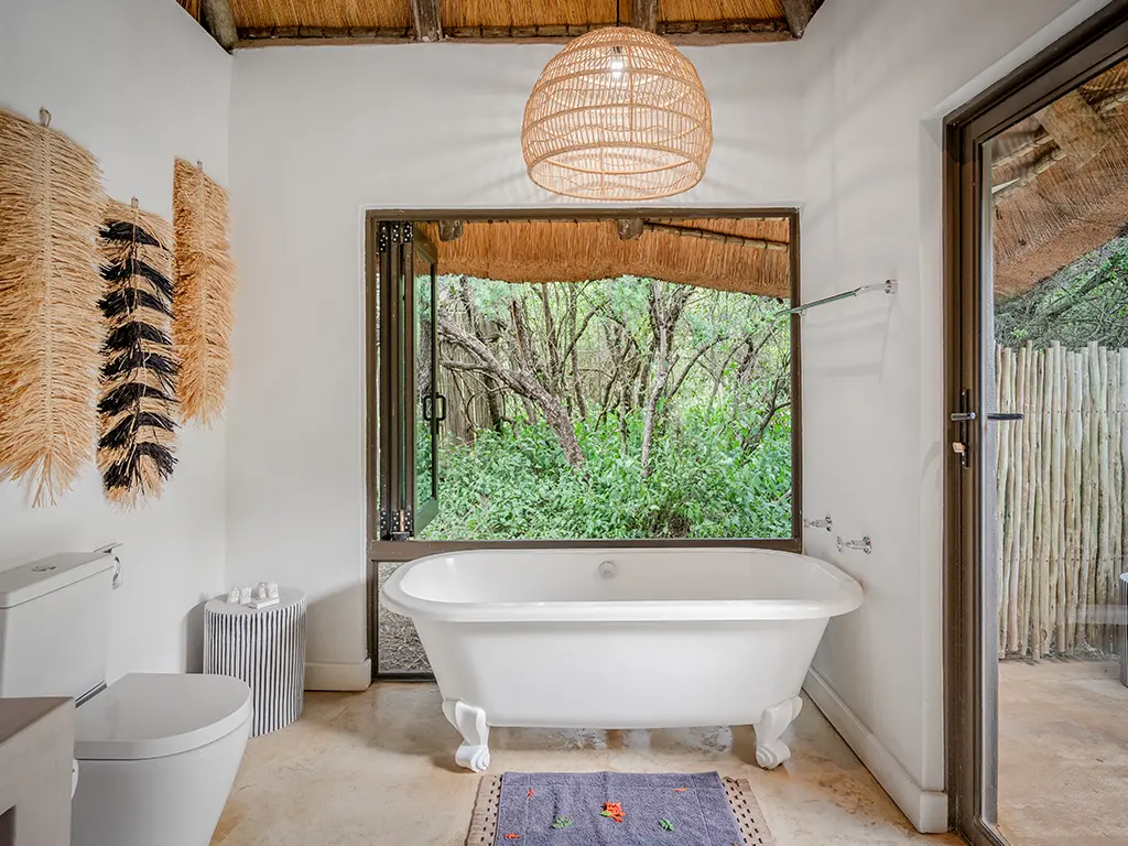 Interior view of a modern en-suite bathroom inside the Homestead at Rhino River Lodge, featuring a white clawfoot bathtub positioned next to a large open window overlooking the green bushveld, a thatched ceiling with a woven pendant light, and a toilet.