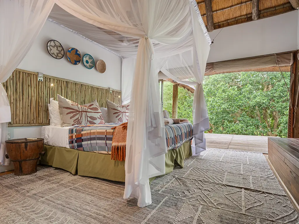 Interior view of another bedroom in the Homestead at Rhino River Lodge, showing a double bed draped in a white mosquito net canopy with a wide textured headboard, next to a large open entrance that leads directly out to the exterior surroundings.