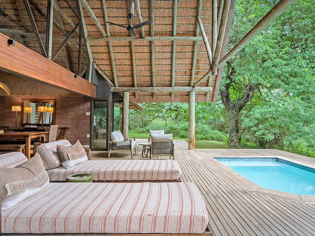 An exterior view of the spacious wooden sun deck outside the Homestead at Rhino River Lodge, featuring comfortable outdoor sofa seating and a large dining table sheltered under a thatched roof structure next to a private swimming pool.