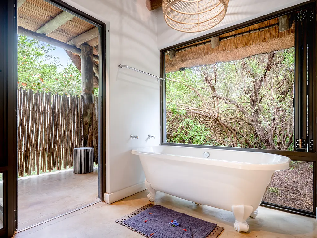 Interior view of another bathroom inside the Homestead at Rhino River Lodge, showing a white bathtub positioned next to a large open window and an exit leading to a private, enclosed outdoor shower area.