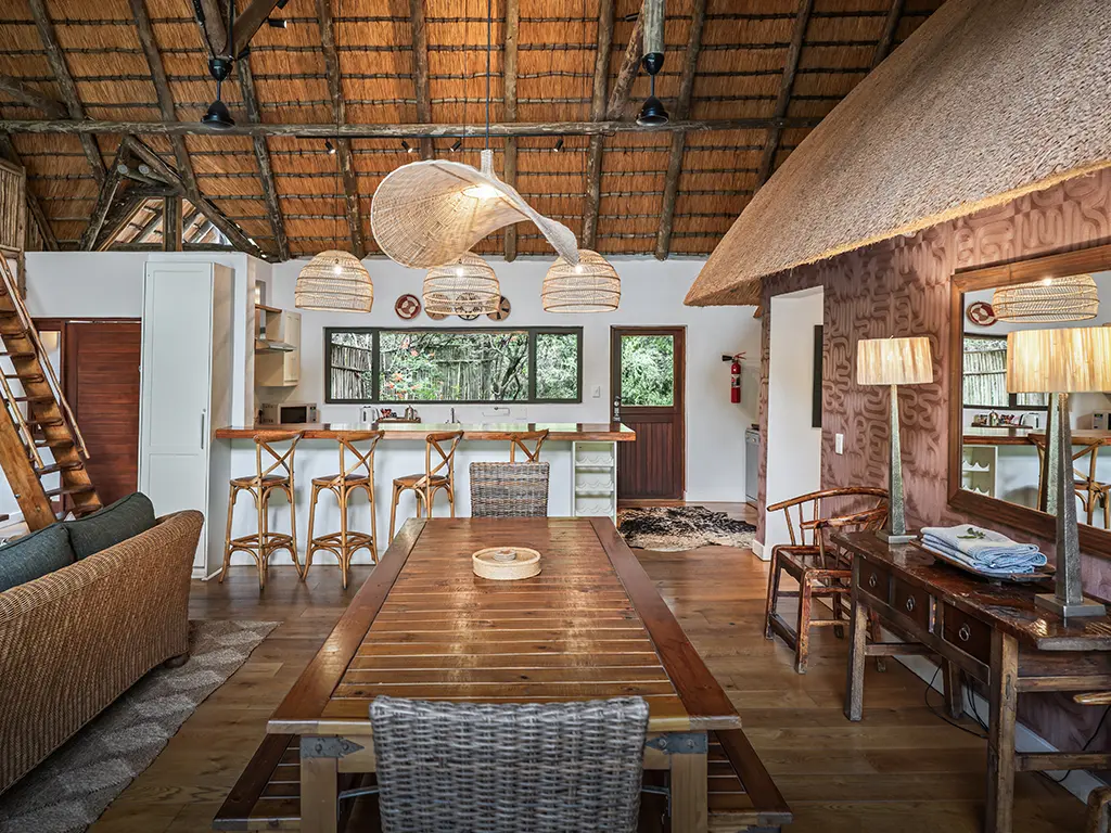 Interior view of the open-plan living and dining area inside the Homestead at Rhino River Lodge, showing a long wooden dining table with chairs set beneath a large woven pendant light, adjacent to a comfortable lounge area with plush sofas and large glass doors leading outside.