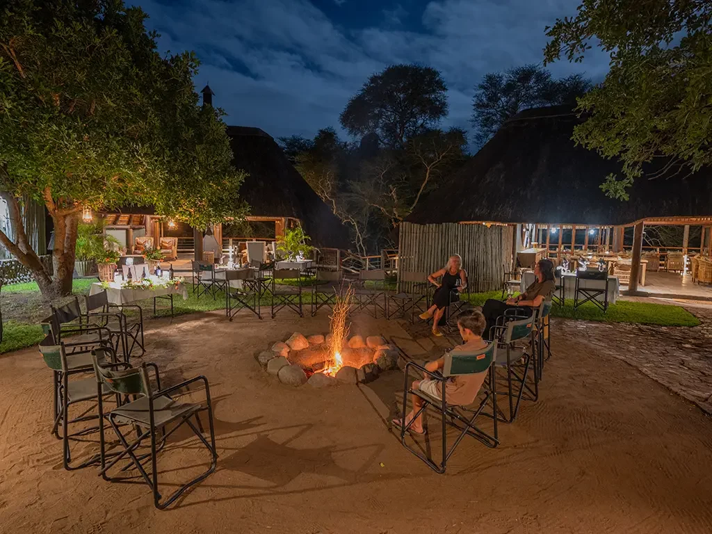 Guests relaxing around a campfire in the open-air boma at night, with the illuminated thatched main buildings of Rhino River Lodge in the background.